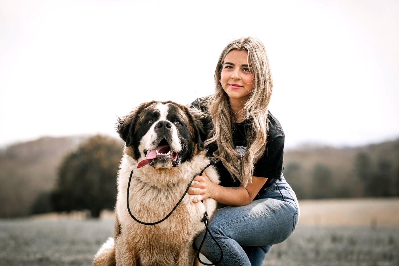 A trainer kneels with her arm around a smiling Saint Bernard in a hilltop field